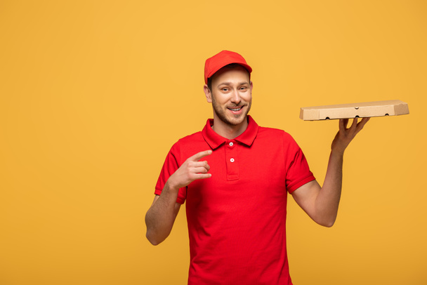 happy delivery man in red uniform pointing with finger at pizza box isolated on yellow - Photo, Image