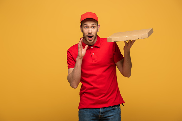 homem de entrega feliz em uniforme vermelho segurando caixa de pizza com boca aberta isolada no amarelo
 - Foto, Imagem