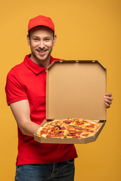 boldog futár piros egyenruhában mutatja doboz finom pizza elszigetelt sárga - Fotó, kép