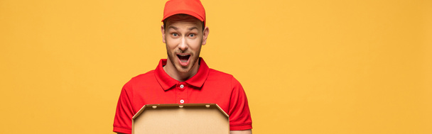 geschokt levering man in rood uniform houden pizza doos geïsoleerd op geel, panoramisch schot - Foto, afbeelding
