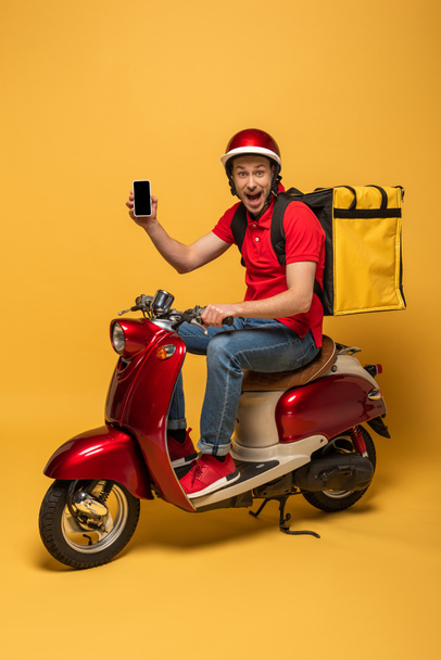 happy delivery man with backpack on scooter holding smartphone on yellow background - Zdjęcie, obraz