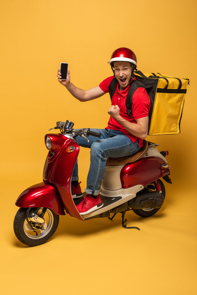 szczęśliwy dostawca z plecakiem na skuterze trzymając smartfona i pokazując tak gest na żółtym tle - Zdjęcie, obraz