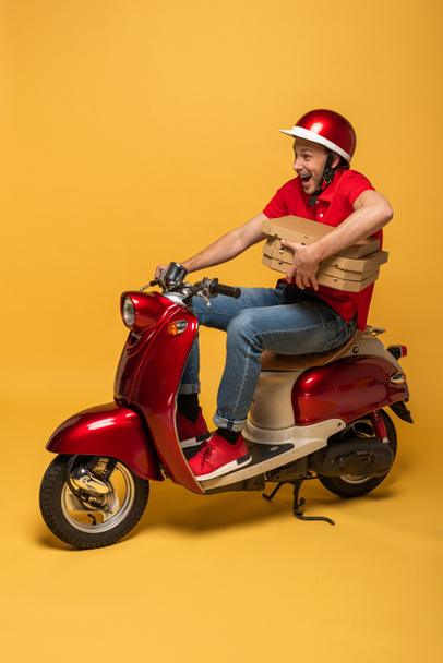 excited delivery man in red uniform holding pizza boxes on scooter on yellow background - Photo, Image