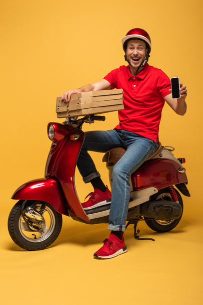 happy delivery man in red uniform holding pizza boxes and smartphone with blank screen on scooter on yellow background - Photo, Image