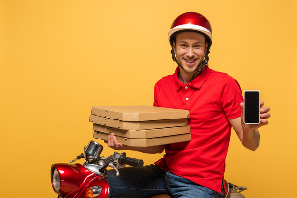 щасливий чоловік доставки в червоній формі тримає коробки для піци і смартфон з порожнім екраном на скутері ізольовані на жовтому
 - Фото, зображення