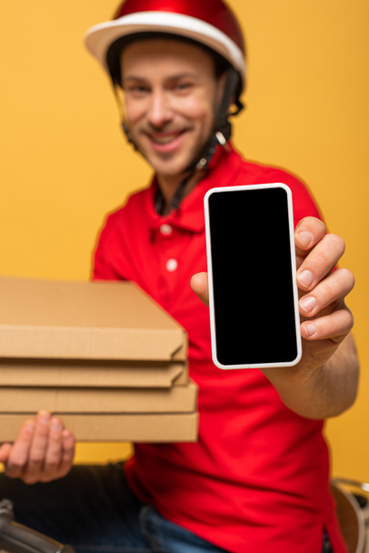 Kırmızı üniformalı mutlu teslimatçının seçici odak noktası pizza kutuları ve sarı ekranda boş ekranlı akıllı telefon. - Fotoğraf, Görsel