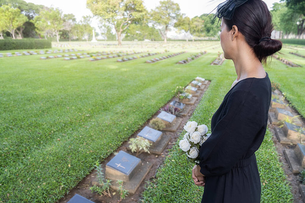 Mourning young woman holding white flowers at her family grave in beautiful green cemetery. - Photo, Image