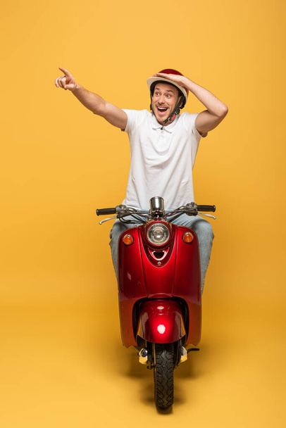 glücklicher Zusteller im Helm, der Roller fährt und mit dem Finger auf gelbem Hintergrund zeigt - Foto, Bild