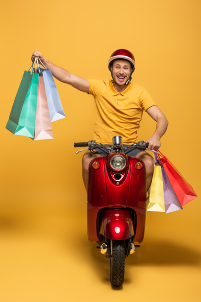happy delivery man in yellow uniform riding scooter with shopping bags on yellow background - Fotografie, Obrázek