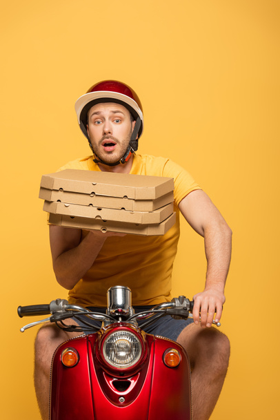 scared delivery man in yellow uniform riding scooter with pizza boxes isolated on yellow - Фото, изображение
