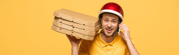 smiling delivery man in yellow uniform with pizza boxes talking on smartphone isolated on yellow, panoramic shot - Valokuva, kuva