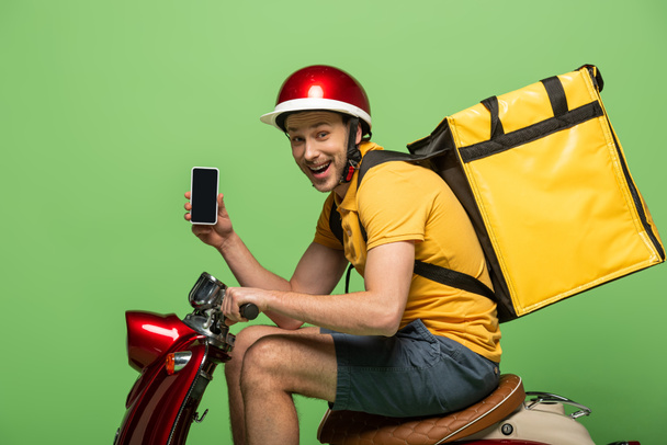 Вид сбоку счастливого доставщика в желтой форме с рюкзаком, показывающим смартфон с экраном на мотороллере, изолированным на зеленом
 - Фото, изображение