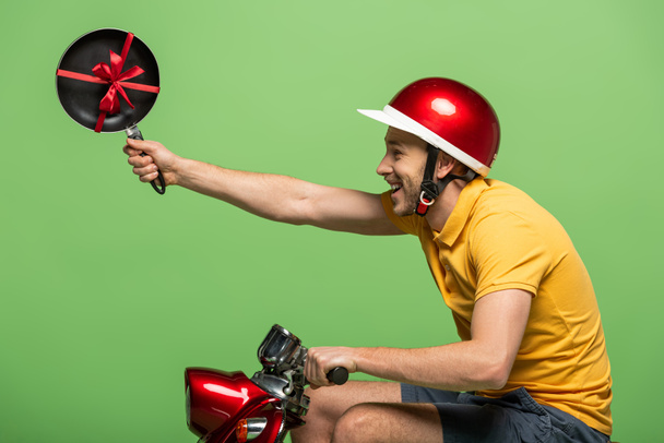 side view of happy delivery man in yellow uniform holding frying pan with bow on scooter isolated on green - Φωτογραφία, εικόνα