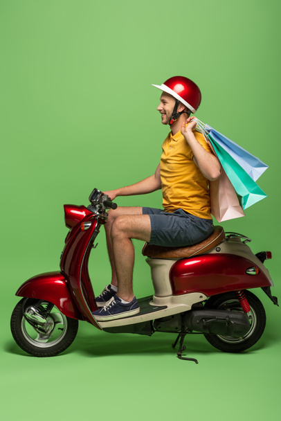 vista lateral do homem de entrega sorridente em uniforme amarelo e capacete segurando sacos de compras em scooter no verde
 - Foto, Imagem