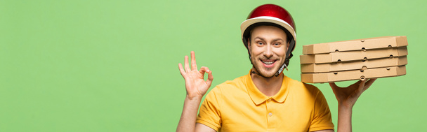 homem de entrega sorrindo em uniforme amarelo entregando pizza e mostrando sinal ok isolado em verde, tiro panorâmico
 - Foto, Imagem
