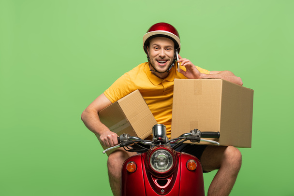 hombre de entrega feliz en uniforme amarillo en scooter con cajas hablando en teléfono inteligente aislado en verde
 - Foto, Imagen