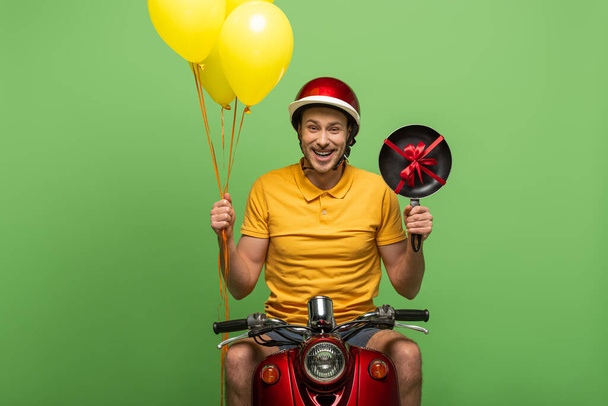 happy delivery man in yellow uniform on scooter with frying pan and balloons isolated on green - Zdjęcie, obraz