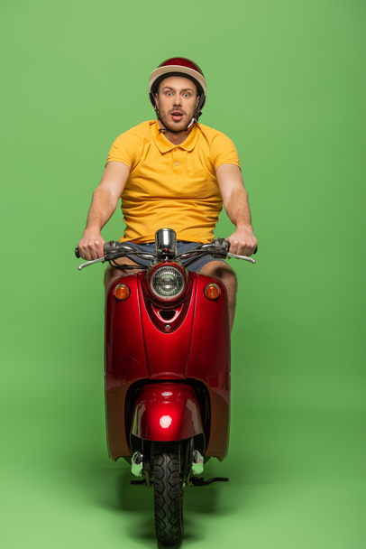shocked delivery man in yellow uniform and helmet on scooter on green - Foto, Bild