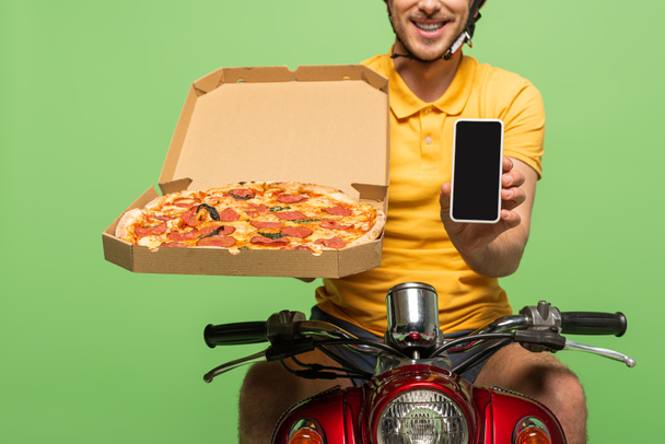 Ausgeschnittene Ansicht eines lächelnden Zustellers in gelber Uniform auf Roller mit Pizza und Smartphone isoliert auf Grün - Foto, Bild