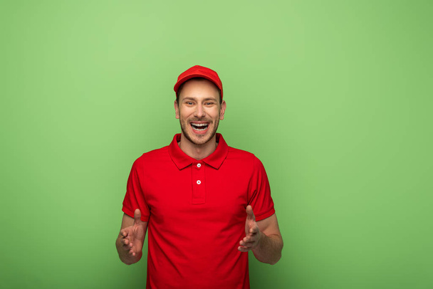 happy delivery man in red uniform gesturing on green - Photo, Image