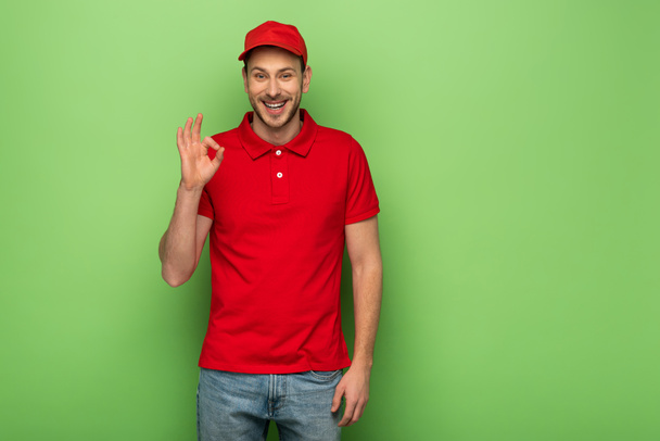 uśmiechnięty dostawca w czerwonym mundurze pokazując ok znak na zielonym tle - Zdjęcie, obraz