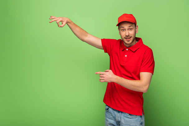 bezorger in rood uniform wijzend met vinger opzij op groene achtergrond - Foto, afbeelding