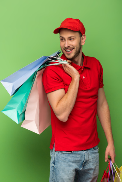 Χαρούμενος ντελιβεράς με κόκκινη στολή που κρατάει τσάντες για ψώνια στο πράσινο - Φωτογραφία, εικόνα
