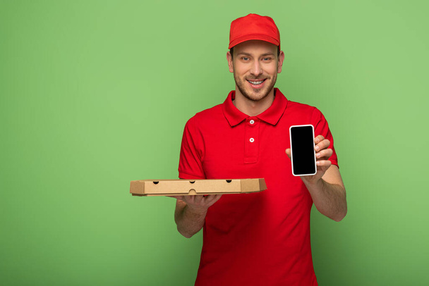 smiling delivery man in red uniform holding pizza box and smartphone on green - Foto, Bild