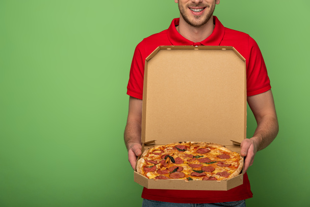 cropped view of smiling delivery man in red uniform holding pizza box on green - Photo, Image
