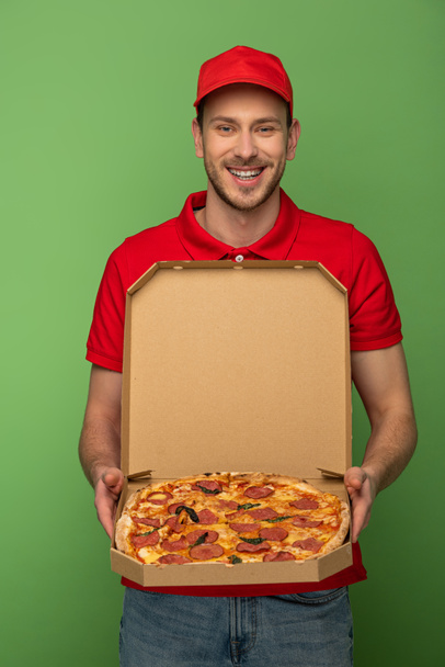 Χαρούμενος ντελιβεράς με κόκκινη στολή κρατώντας κουτί πίτσας στο πράσινο - Φωτογραφία, εικόνα