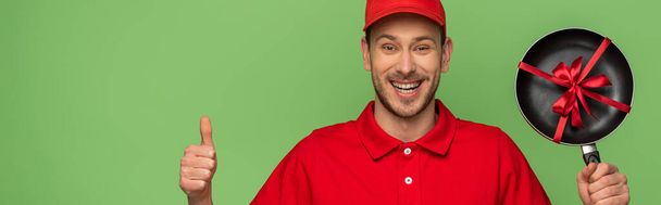 happy delivery man in red uniform holding frying pan with bow and showing thumb up isolated on green - Foto, afbeelding