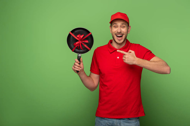 happy delivery man in red uniform pointing with finger at frying pan with bow on green - Foto, afbeelding