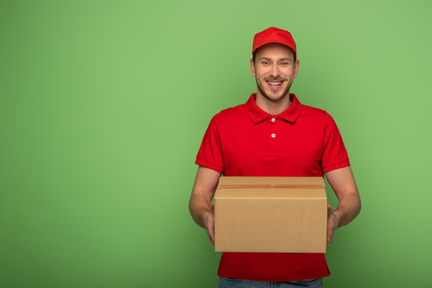 усміхнений чоловік доставки в червоній формі тримає посилку на зеленому
 - Фото, зображення