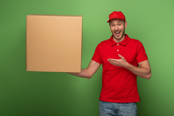 onnellinen toimitus mies punainen univormu osoittaa käsi valtava paketti vihreä
 - Valokuva, kuva