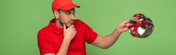 pensive delivery man in red uniform holding pan with bow isolated on green, panoramic shot - Фото, изображение