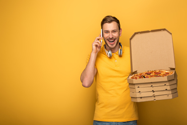 happy man with headphones in yellow outfit holding pizza and talking on smartphone on yellow background - Photo, Image