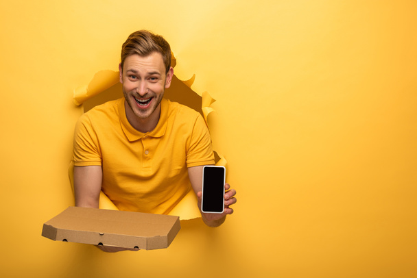happy handsome man in yellow outfit holding smartphone and pizza box in yellow paper hole  - Photo, Image