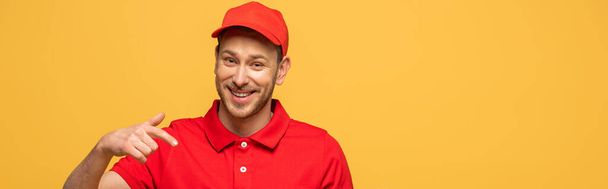 happy delivery man in red uniform pointing with finger isolated on yellow, panoramic shot - Foto, immagini