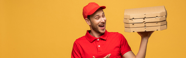 onnellinen toimitus mies punainen univormu osoittaa käsi kohti pizza laatikot eristetty keltainen, panoraama laukaus
 - Valokuva, kuva