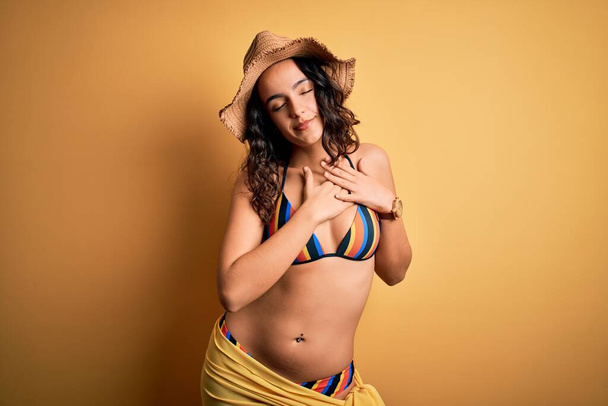 Young beautiful woman with curly hair on vacation wearing bikini and summer hat smiling with hands on chest with closed eyes and grateful gesture on face. Health concept. - 写真・画像