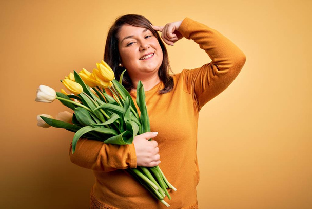 Krásná plus velikost žena drží romantickou kytici přírodních tulipánů květy na žlutém pozadí Usmívající se ukazuje na hlavu s jedním prstem, skvělý nápad nebo myšlenka, dobrá paměť - Fotografie, Obrázek