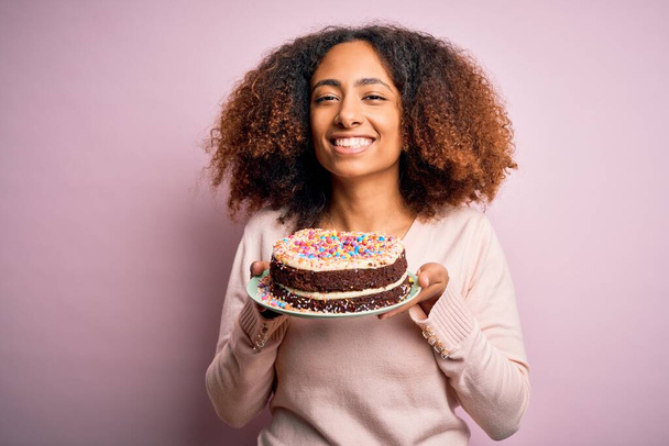 Jonge Afro-Amerikaanse vrouw met afro haar houdt heerlijke verjaardagstaart met een vrolijk gezicht staande en glimlachend met een zelfverzekerde glimlach tonen tanden - Foto, afbeelding