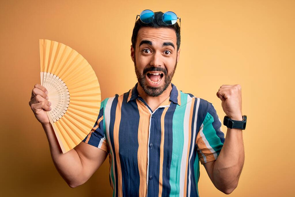Νεαρός όμορφος τουρίστας με γενειάδα σε διακοπές φορώντας καλοκαιρινό πουκάμισο χρησιμοποιώντας το χέρι ανεμιστήρα ουρλιάζοντας υπερήφανος και γιορτάζει τη νίκη και την επιτυχία πολύ ενθουσιασμένος, ζητωκραυγάζοντας συναίσθημα - Φωτογραφία, εικόνα