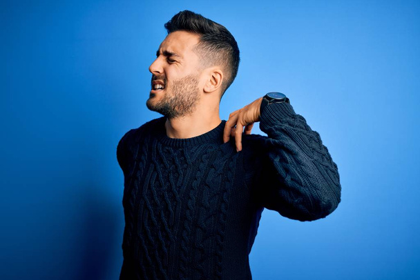 Young handsome man wearing casual sweater standing over isolated blue background Suffering of neck ache injury, touching neck with hand, muscular pain - Fotografie, Obrázek