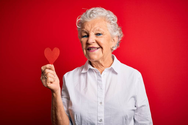 Senior bela mulher segurando papel coração de pé sobre fundo vermelho isolado com um rosto feliz de pé e sorrindo com um sorriso confiante mostrando dentes
 - Foto, Imagem