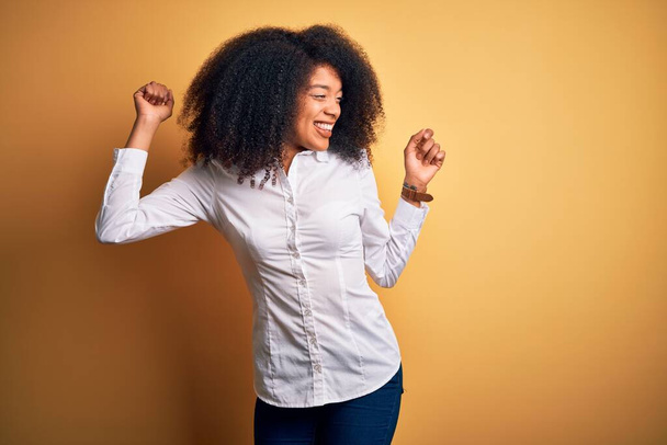 Молода красива афроамериканська елегантна жінка з афро волоссям, що стоїть над жовтим тлом Танці щасливі і веселі, посміхаючись рухаючись випадково і впевнено слухаючи музику
 - Фото, зображення