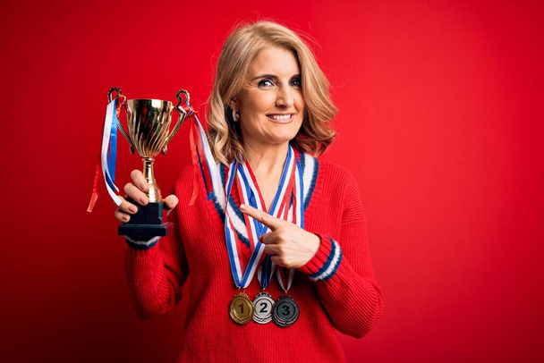Middle age beautiful blonde successful woman wearing medals holding trophy very happy pointing with hand and finger - Valokuva, kuva