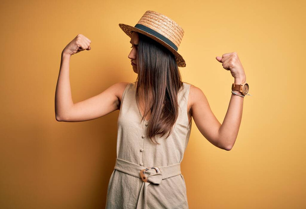 Joven mujer morena hermosa de vacaciones con vestido casual y sombrero mostrando los músculos de los brazos sonriendo orgulloso. Concepto Fitness
. - Foto, imagen