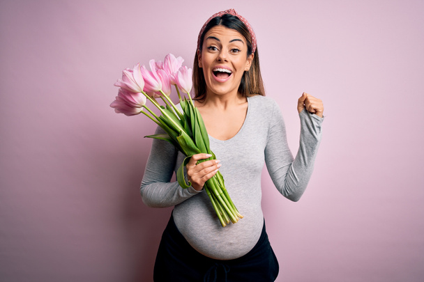 Νέα όμορφη μελαχρινή γυναίκα έγκυος περιμένει μωρό κρατώντας μπουκέτο τουλίπες ουρλιάζοντας υπερήφανοι και γιορτάζει τη νίκη και την επιτυχία πολύ ενθουσιασμένος, ζητωκραυγάζοντας συναίσθημα - Φωτογραφία, εικόνα