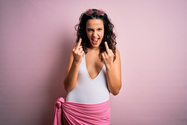 Beautiful woman with curly hair on vacation wearing white swimsuit over pink background Showing middle finger doing fuck you bad expression, provocation and rude attitude. Screaming excited - Photo, Image